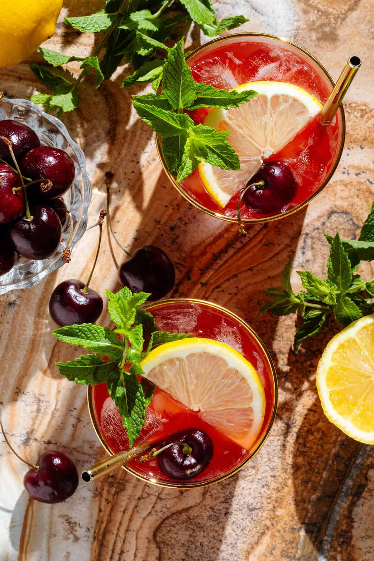 A red bourbon cherry cocktail in two short glasses with a gold rim garnished with a slices lemon, fresh cherries, and fresh mint with more garnish around the glasses.
