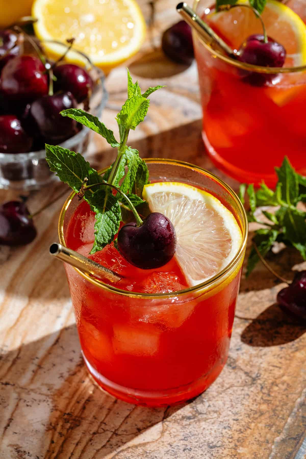 A red bourbon cherry cocktail in two short glasses with a gold rim garnished with a slices lemon, fresh cherries, and fresh mint.