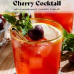 A red bourbon cherry cocktail in a short glass with a gold rim garnished with a slice of lemon, a fresh cherry, and a sprig of fresh mint.