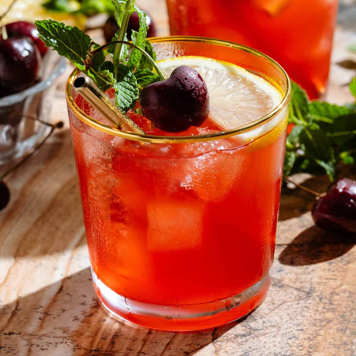 A red bourbon cherry cocktail in a short glass with a gold rim garnished with a slice of lemon, a fresh cherry, and a sprig of fresh mint.