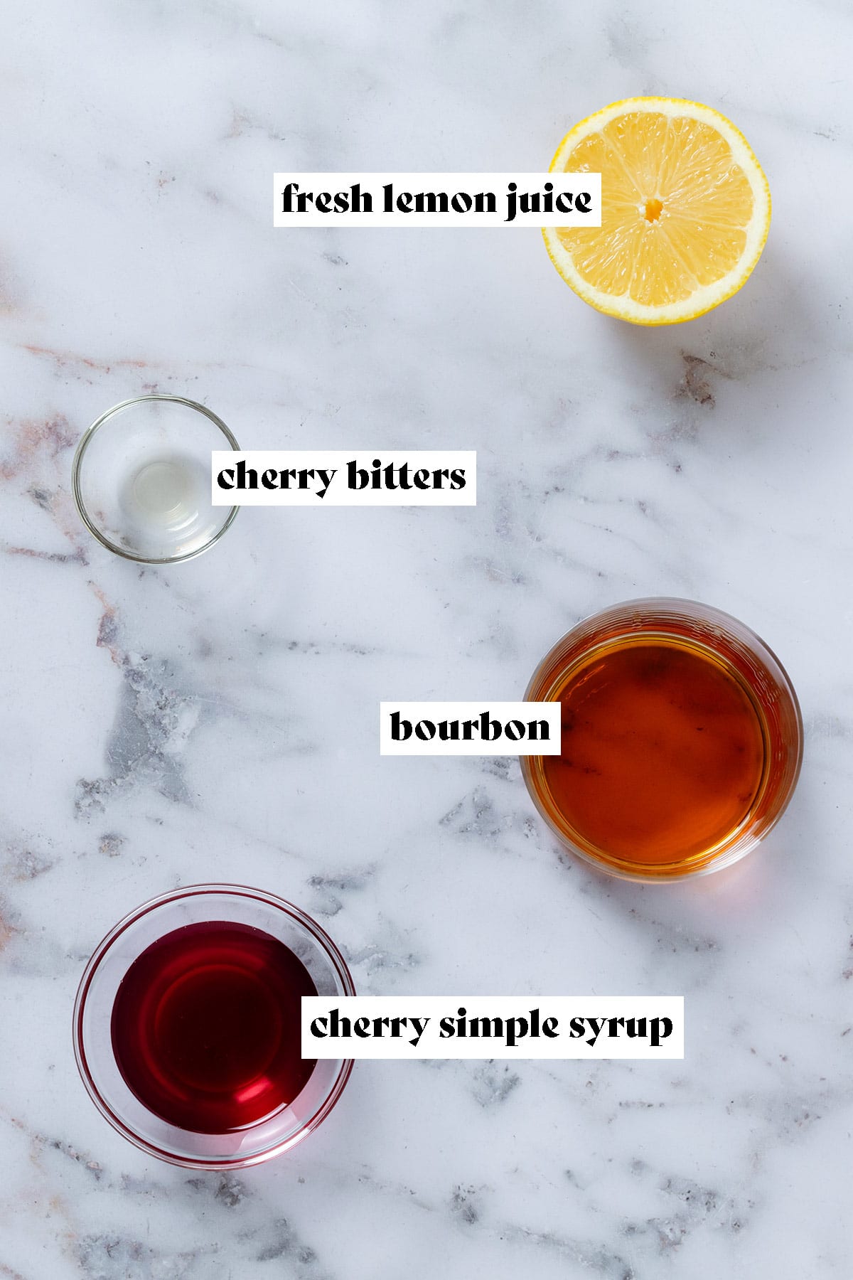 Ingredients like bourbon, cherry syrup, and bitters laid out in small glass bowls, and half a lemon in the top right.