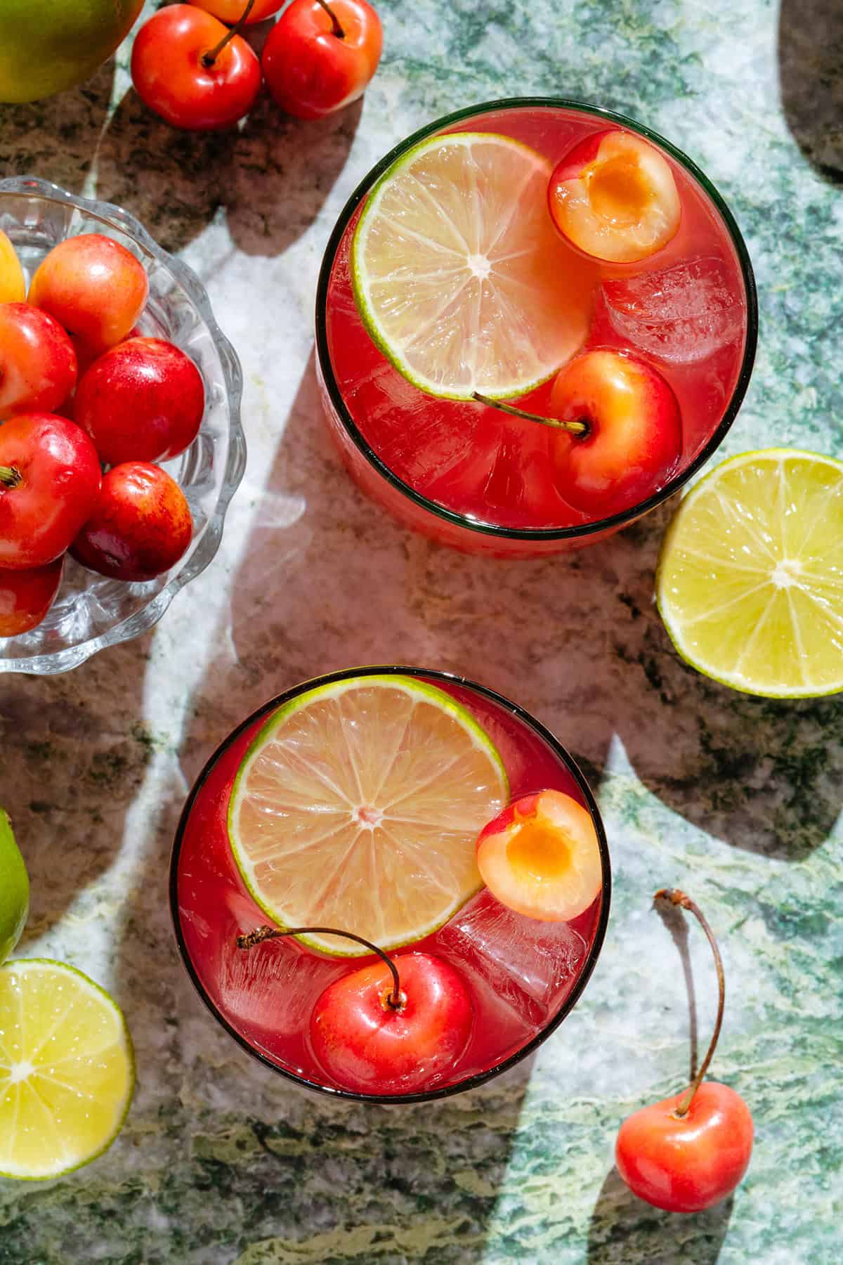 Bright red cherry margarita in two short glasses garnished with slices of lime and fresh cherries.