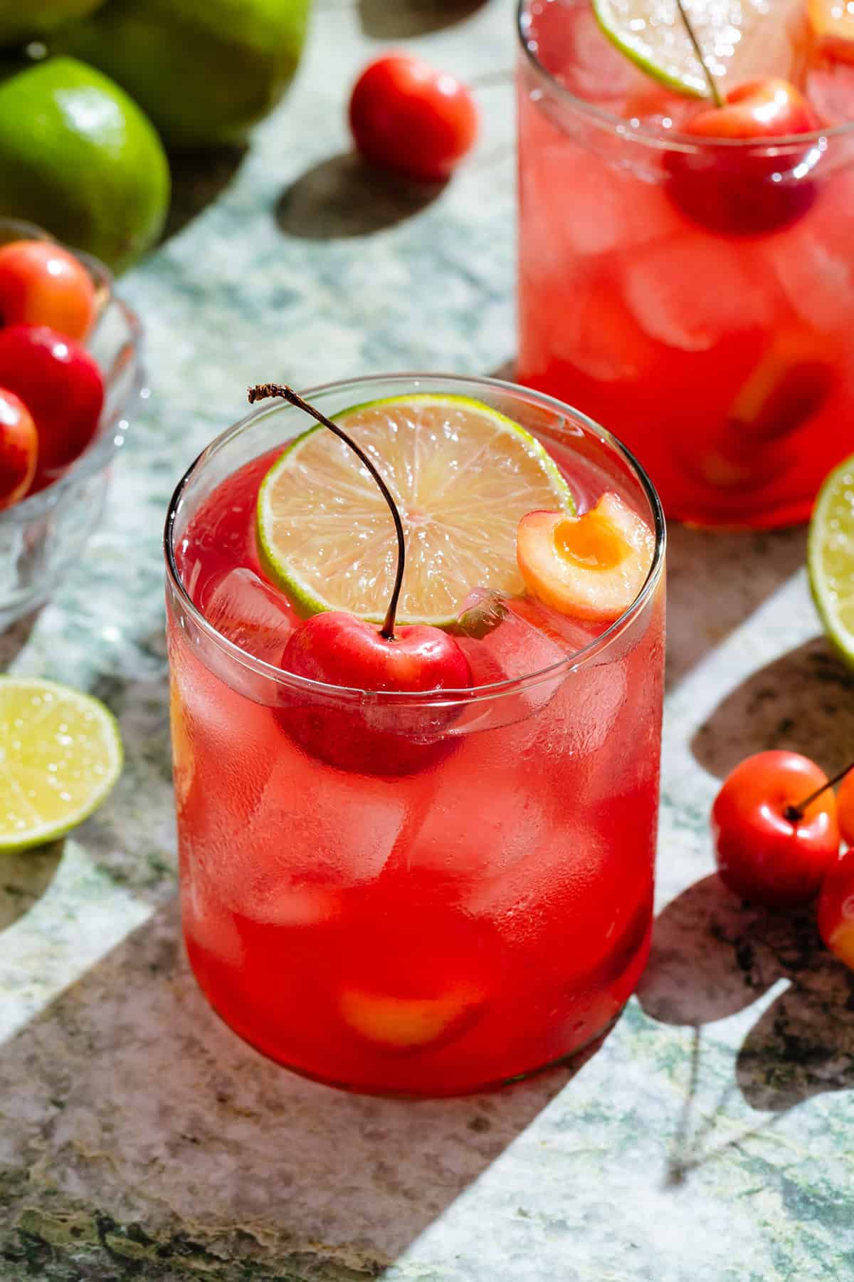 Bright red cherry margarita in two short glasses garnished with slices of lime and fresh cherries.