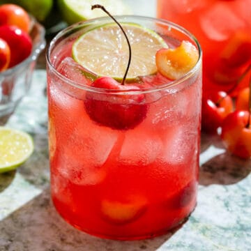 Bright red cherry margarita in a short glass garnished with a slice of lime and fresh cherries.