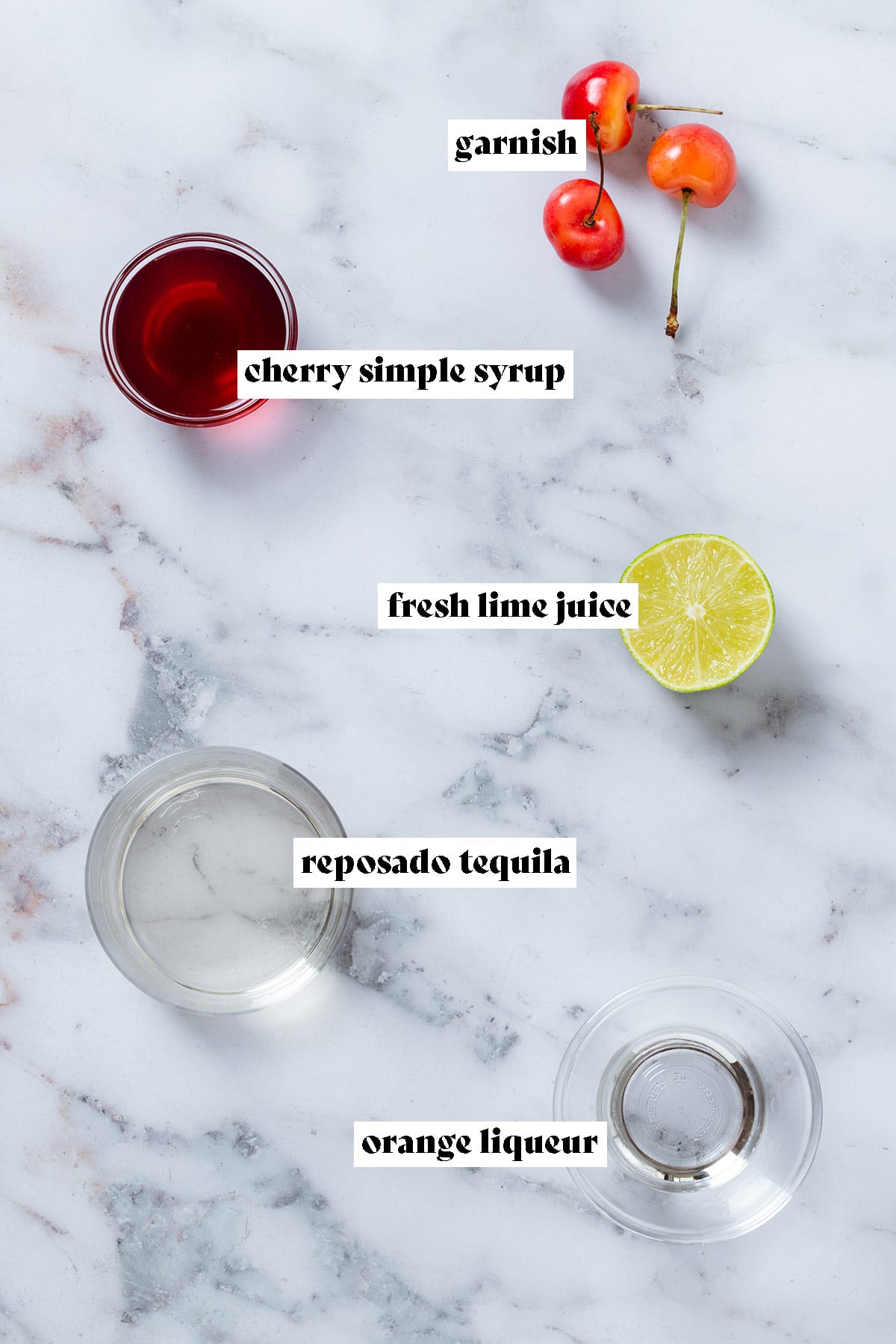 Ingredients like cherry syrup, tequila, and orange liqueur in small glass bowls with fresh cherries and half of a lime next to them.
