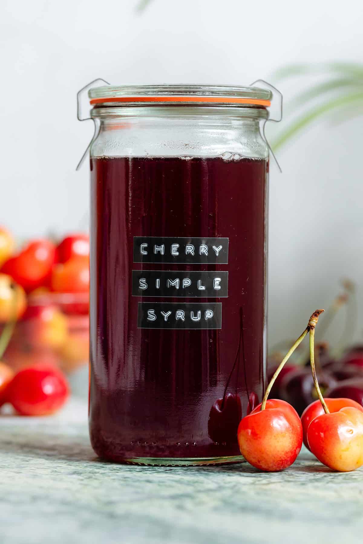 A tall glass jar with dark red cherry syrup with light and dark cherries all around it.
