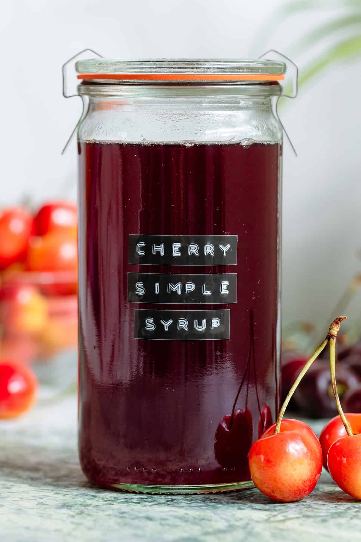 A tall glass jar with dark red cherry syrup with light and dark cherries all around it.