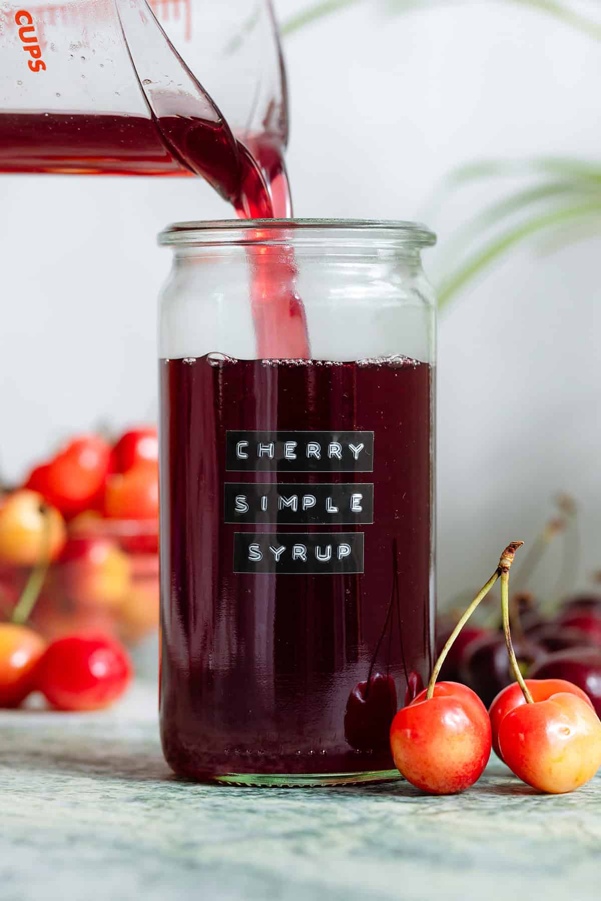 Dark red cherry syrup being poured into a tall glass jar with cherries all around it.