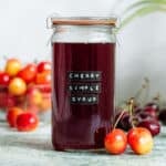 A glass jar with dark red cherry syrup with light and dark cherries all around it.