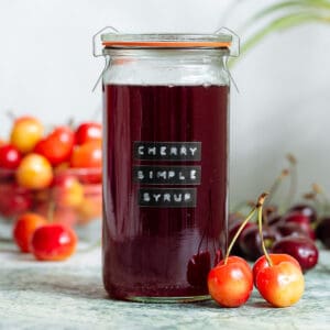 A glass jar with dark red cherry syrup with light and dark cherries all around it.