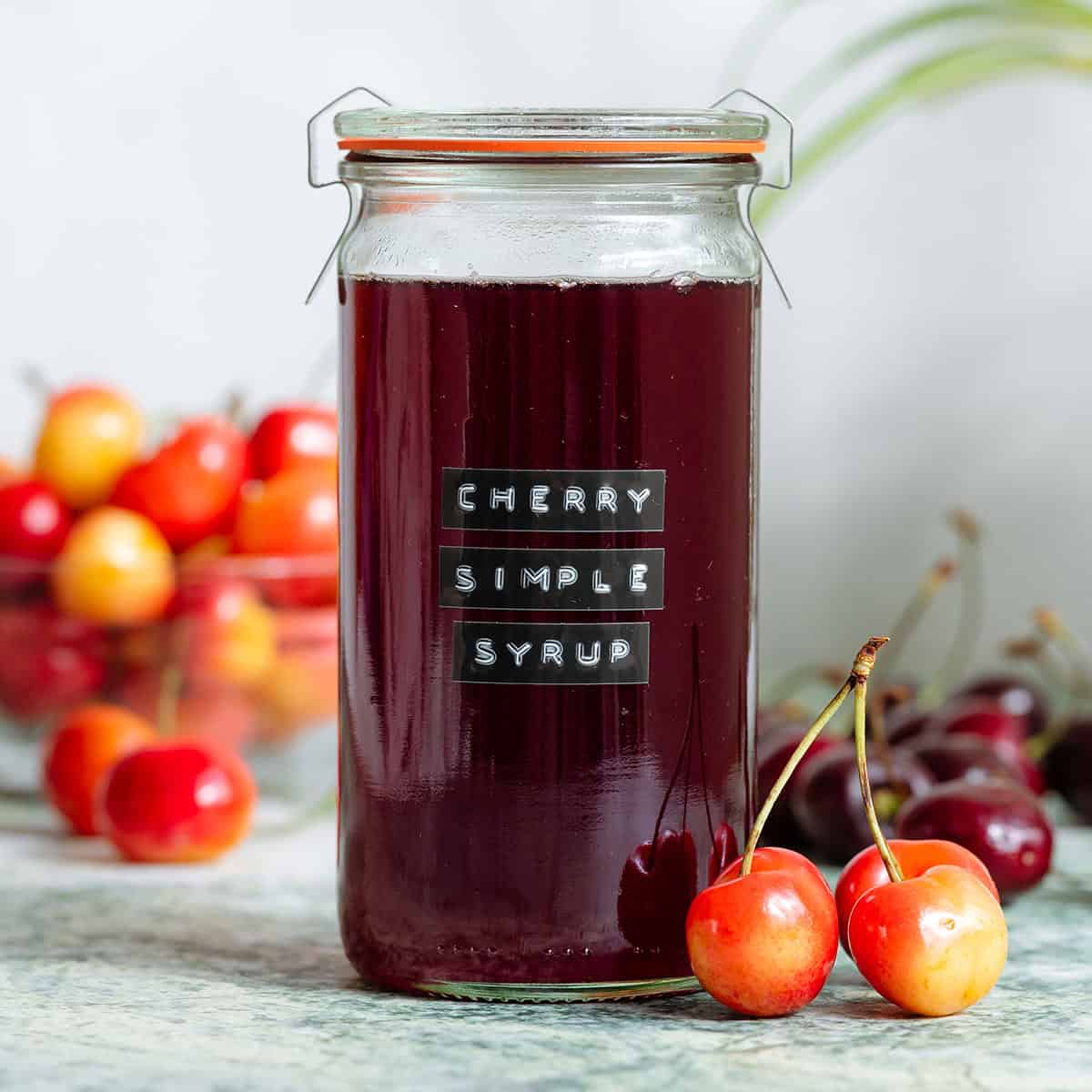 A glass jar with dark red cherry syrup with light and dark cherries all around it.