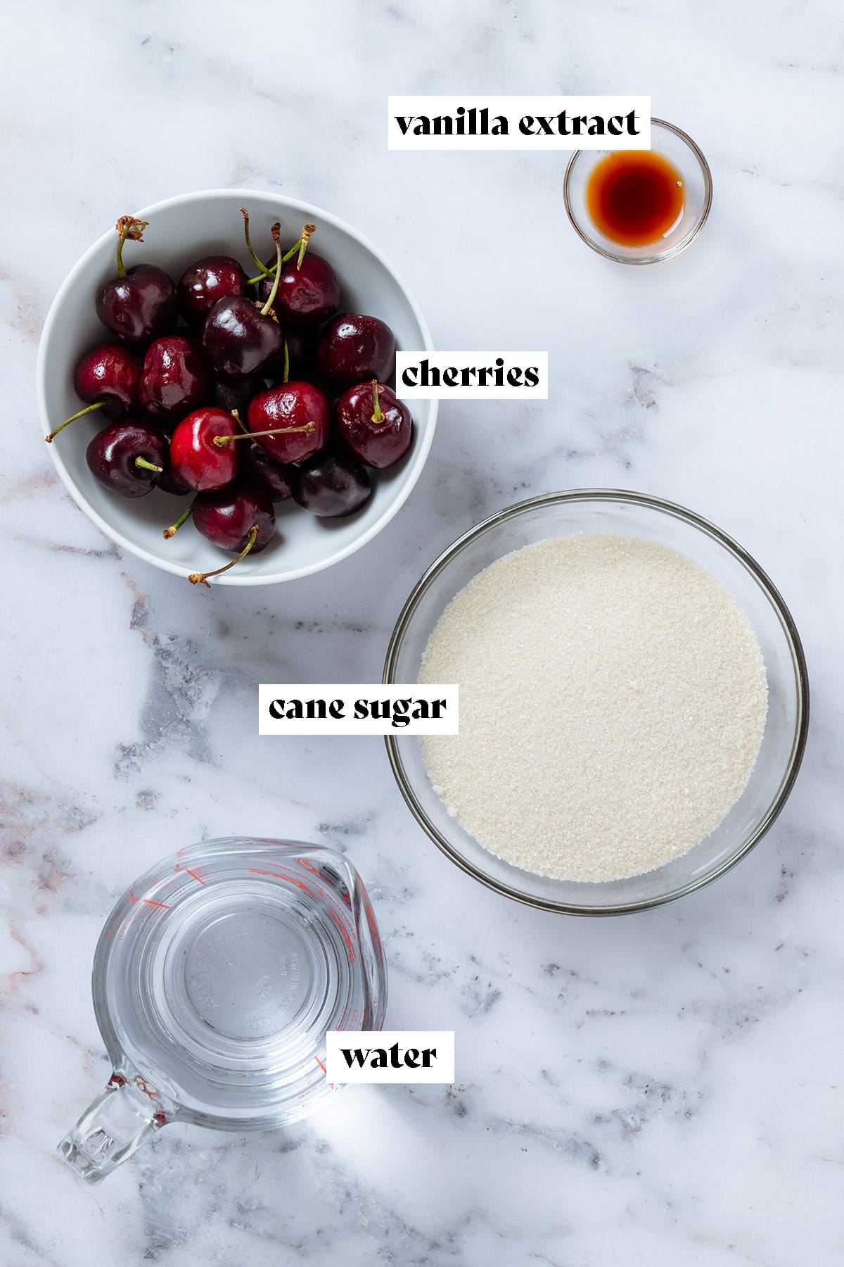 Fresh cherries and cane sugar in bowls, water in a measuring cup, and vanilla extract in tiny bowl laid out.