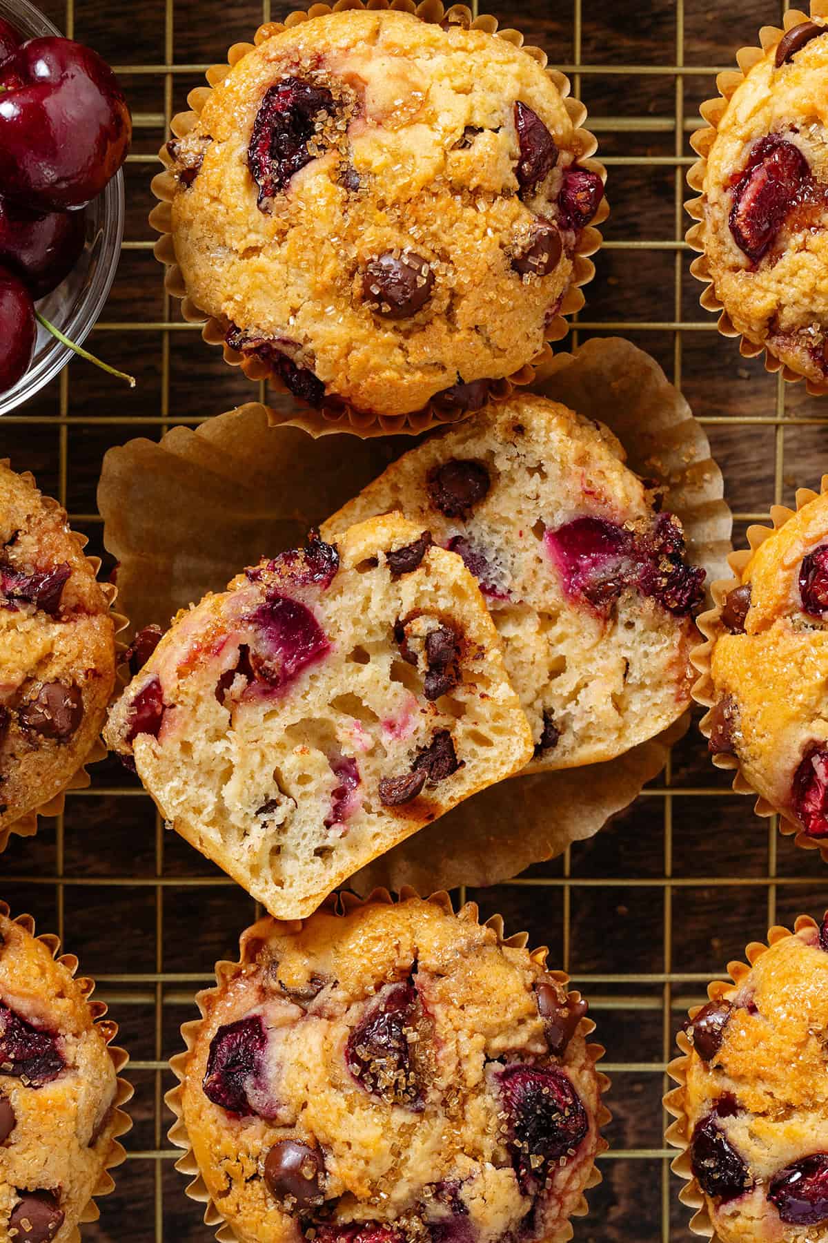 Muffins with cherries and chocolate chips on a cooling rack with the middle one cut in half to show the fluffy inside.