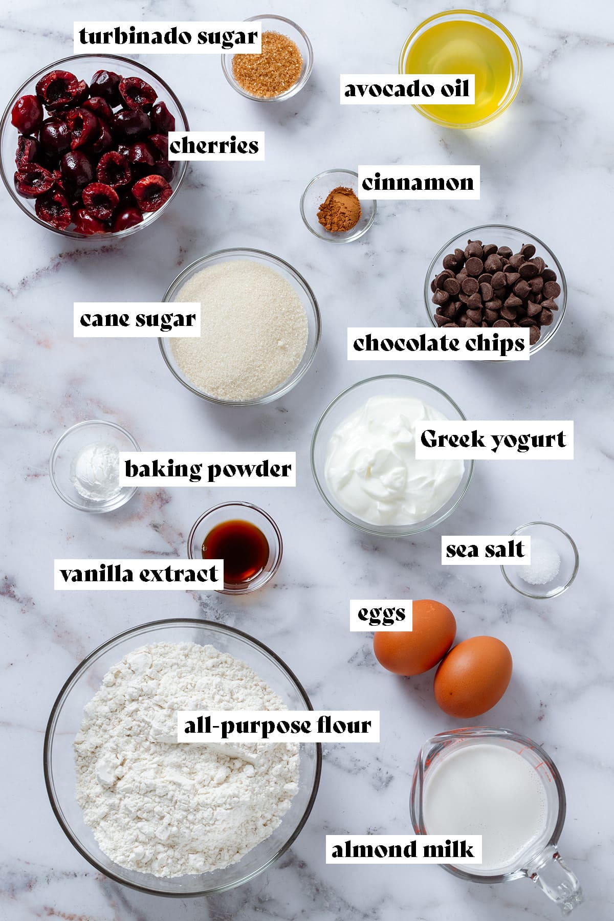 Ingredients like flour, fresh cherries, eggs, chocolate chips, cane sugar, yogurt, oil, and baking powder in small glass bowls on marble background.