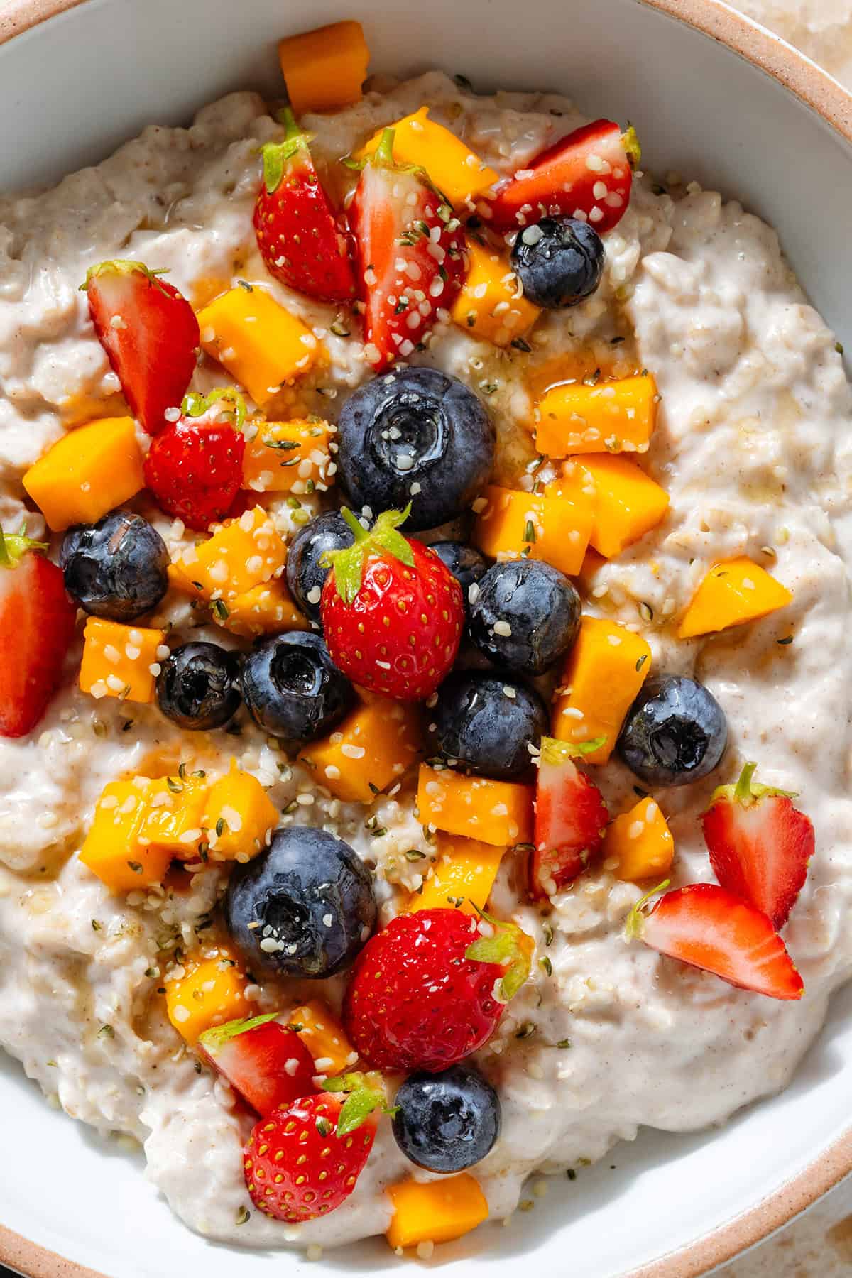 Creamy yogurt oatmeal in a white bowl with a beige rim topped with blueberries, strawberries, chopped mango, and drizzled with honey.