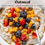 Creamy yogurt oatmeal in a white bowl with a beige rim topped with blueberries, strawberries, chopped mango, and drizzled with honey with a spoon.