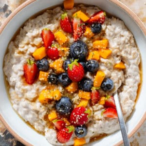 Creamy yogurt oatmeal in a white bowl with a beige rim topped with blueberries, strawberries, chopped mango, and drizzled with honey.