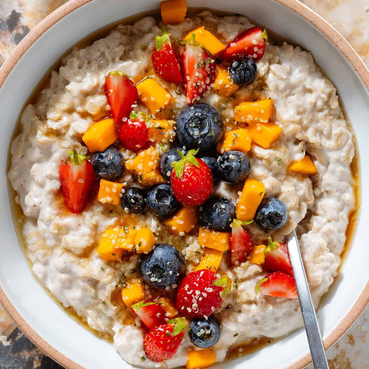 Creamy yogurt oatmeal in a white bowl with a beige rim topped with blueberries, strawberries, chopped mango, and drizzled with honey.