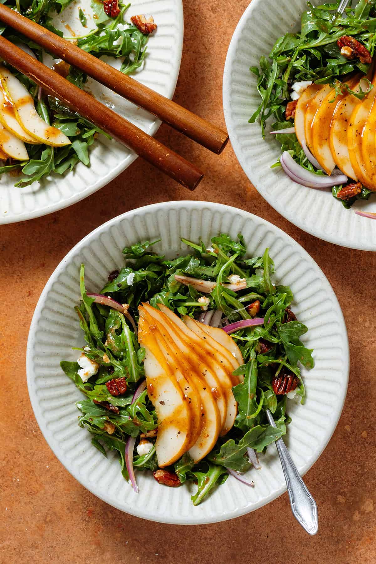 Arugula salad with sliced pears, feta, and roasted chopped pecans drizzled with balsamic dressing in two low bowls with forks.