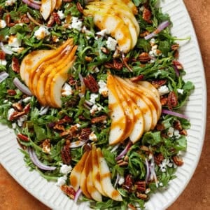 Arugula salad with sliced pears, feta, and roasted chopped pecans on a large serving plate drizzled with balsamic dressing.