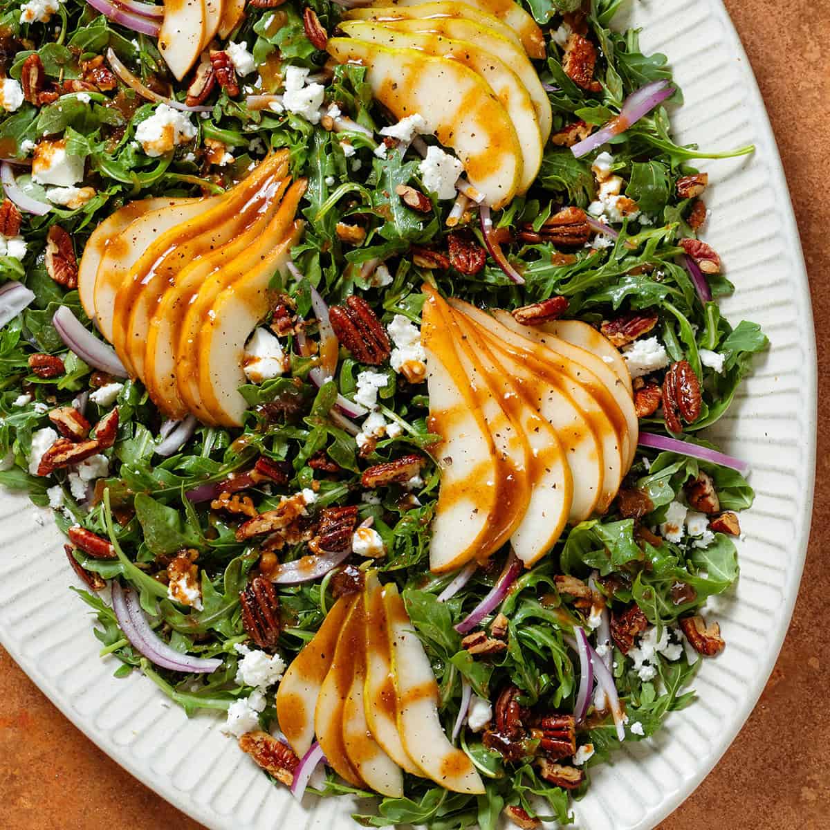 Arugula salad with sliced pears, feta, and roasted chopped pecans on a large serving plate drizzled with balsamic dressing.