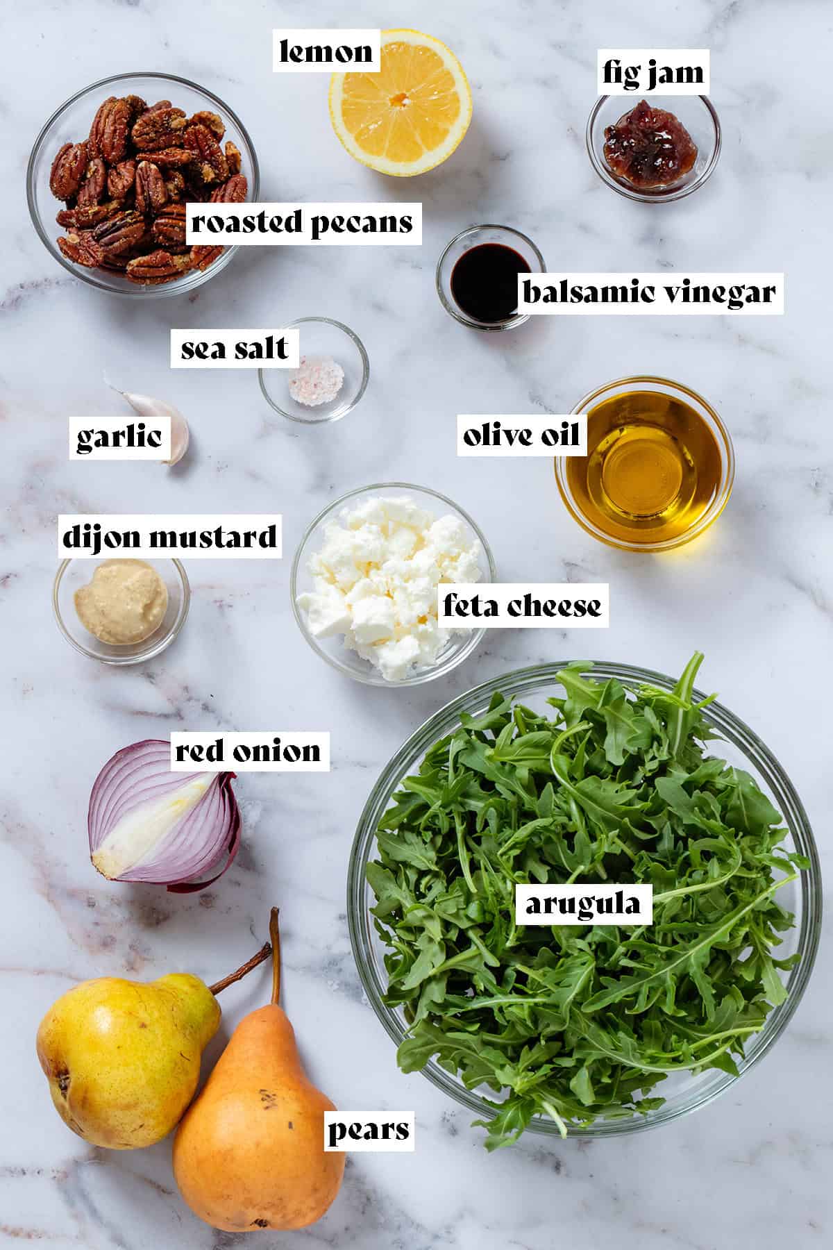 Ingredients in small glass bowls like feta, roasted pecans, olive oil, dijon mustard, and arugula.