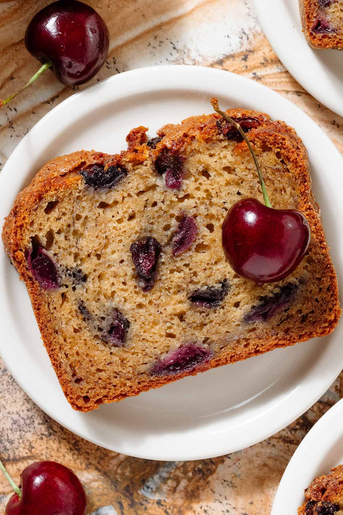 A slice of banana bread with cherries on a small white plate.