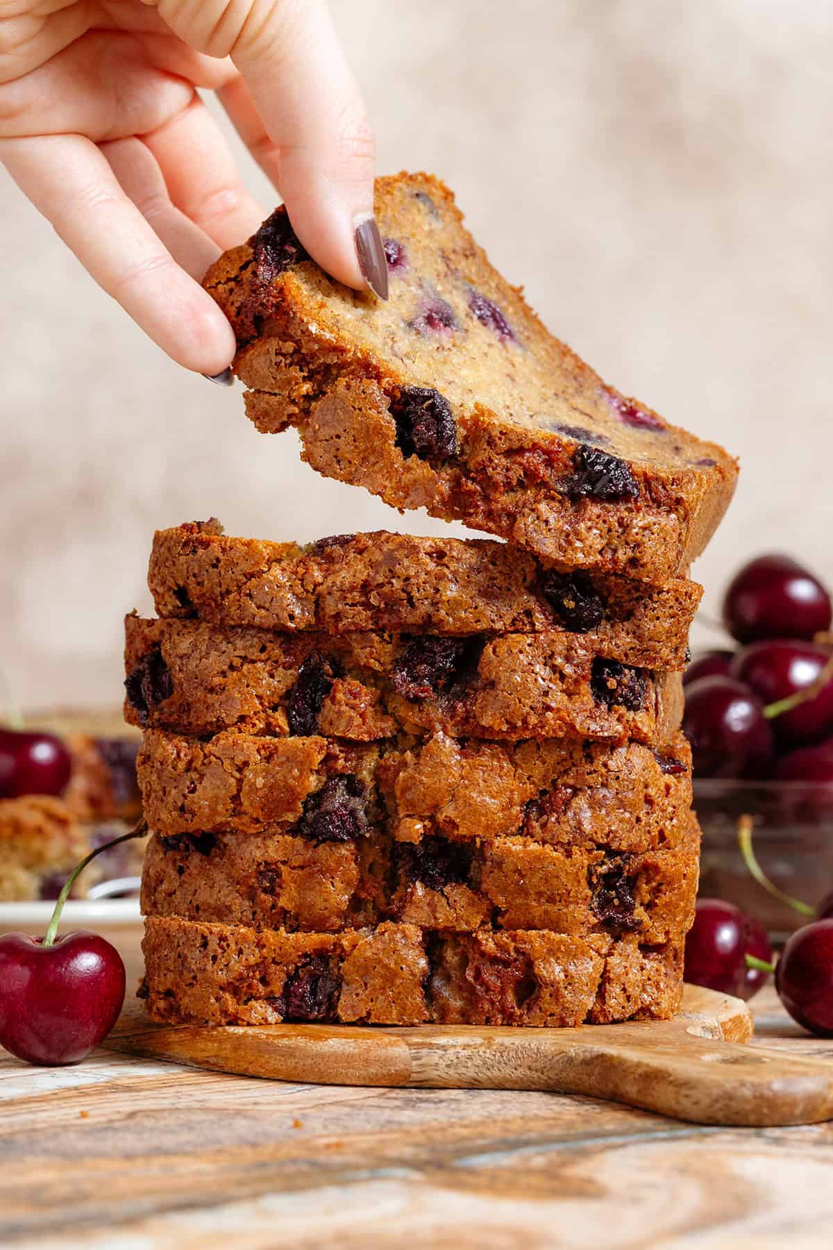 A stack of cherry banana bread slices on a small cutting board and a hand taking a slice off the top.