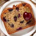 A slice of banana bread with cherries on a small white plate.