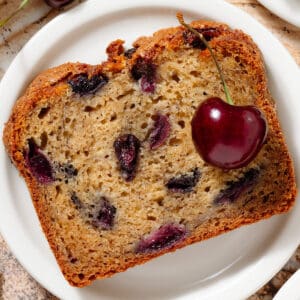 A slice of banana bread with cherries on a small white plate.