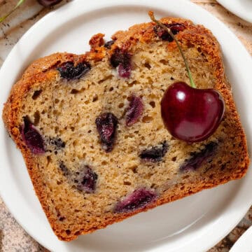 A slice of banana bread with cherries on a small white plate.