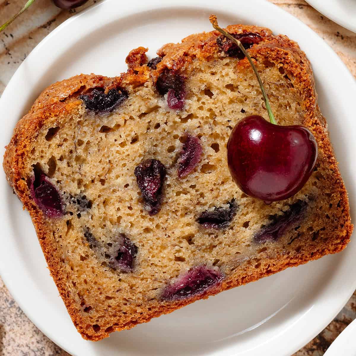 A slice of banana bread with cherries on a small white plate.