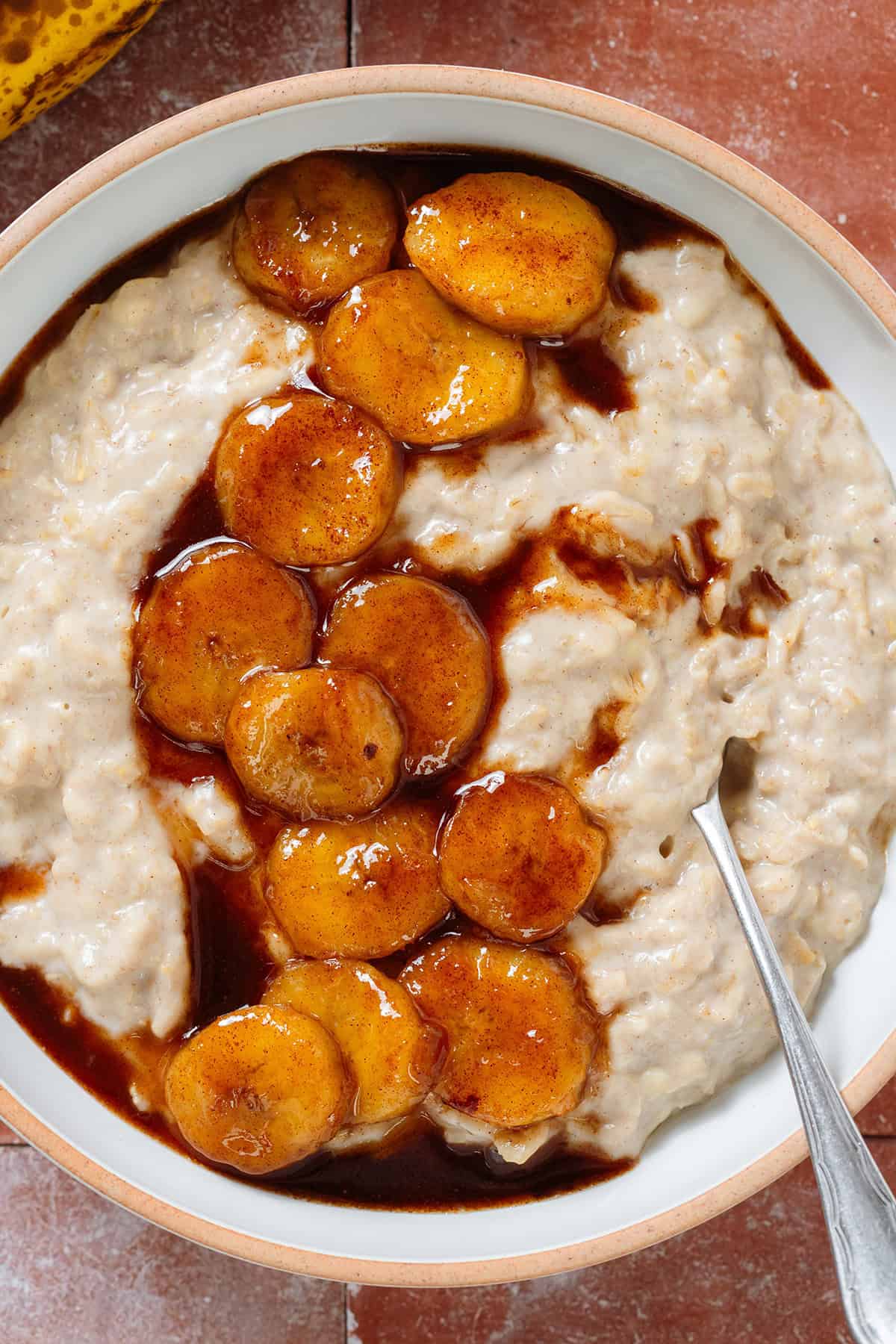 Creamy oatmeal in a white bowl topped with caramelized banana slices with lots of thick sauce from cooking in maple syrup, and with a spoon in the bowl on the right.