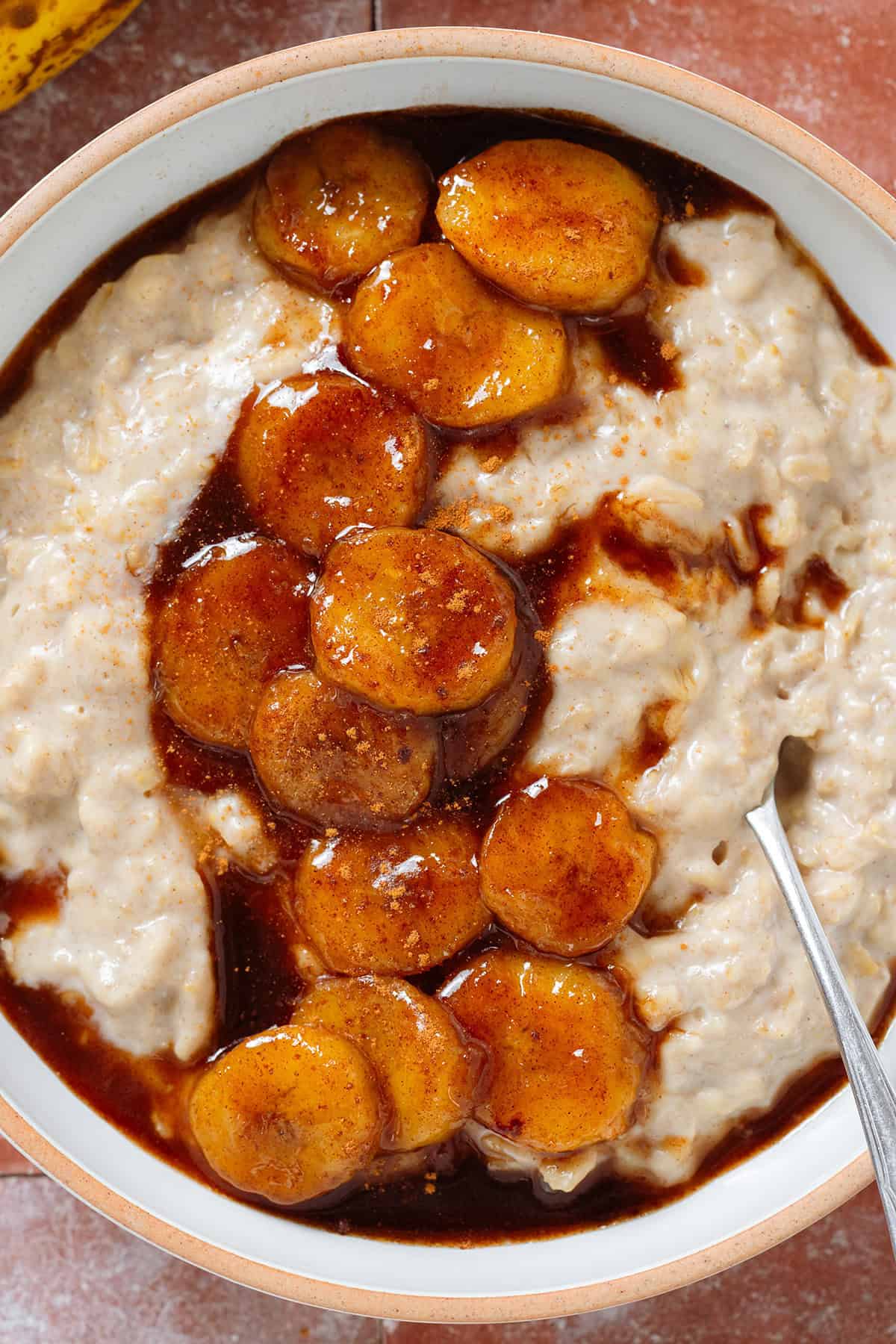 Creamy oatmeal in a white bowl topped with caramelized banana slices cooked with maple syrup, sprinkled with cinnamon, and with a spoon in the bowl on the right.