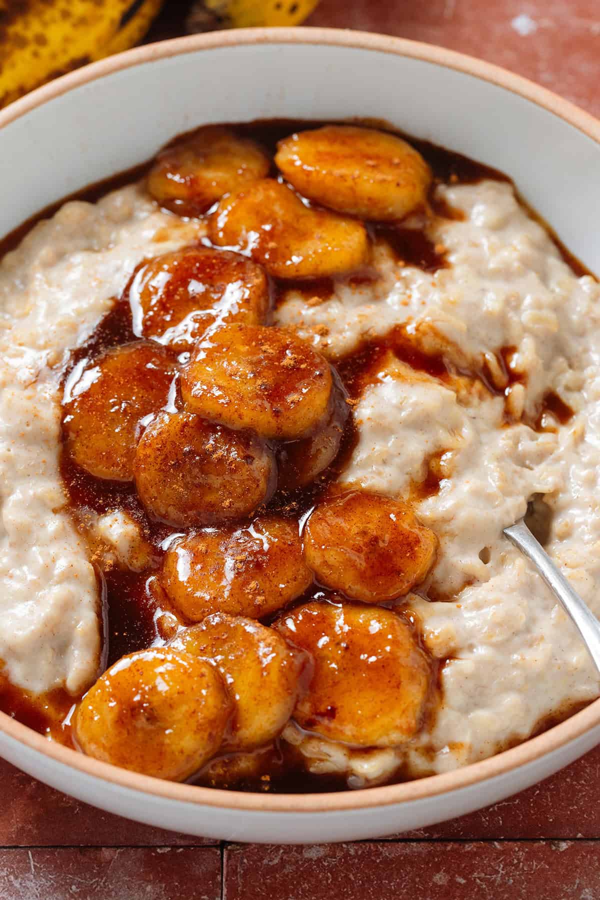 Creamy oatmeal in a white bowl topped with caramelized banana slices cooked with maple syrup, sprinkled with cinnamon, and with a spoon in the bowl on the right.