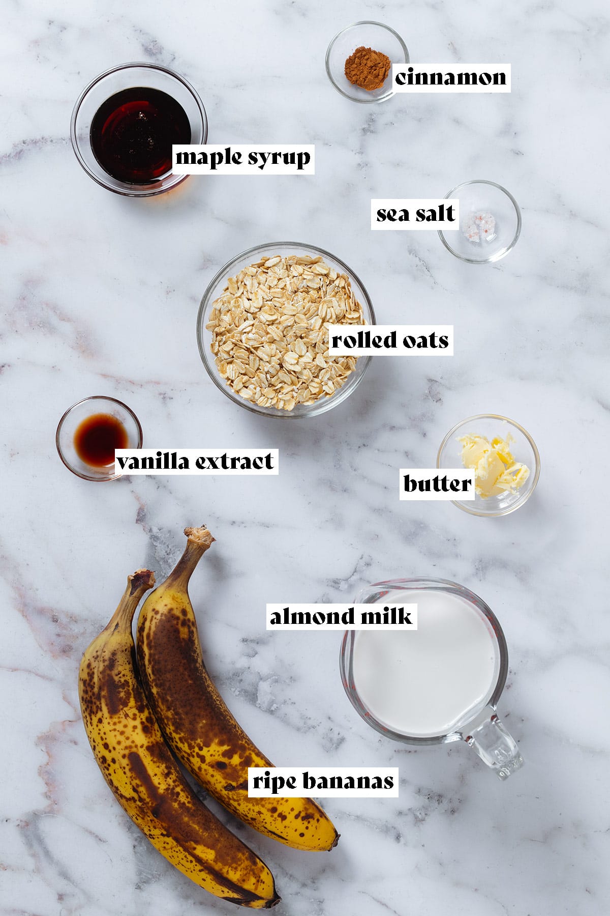Ingredients like rolled oats, butter, maple syrup, vanilla extract in small bowls, and two ripe bananas laid out on marble background with text overlay.
