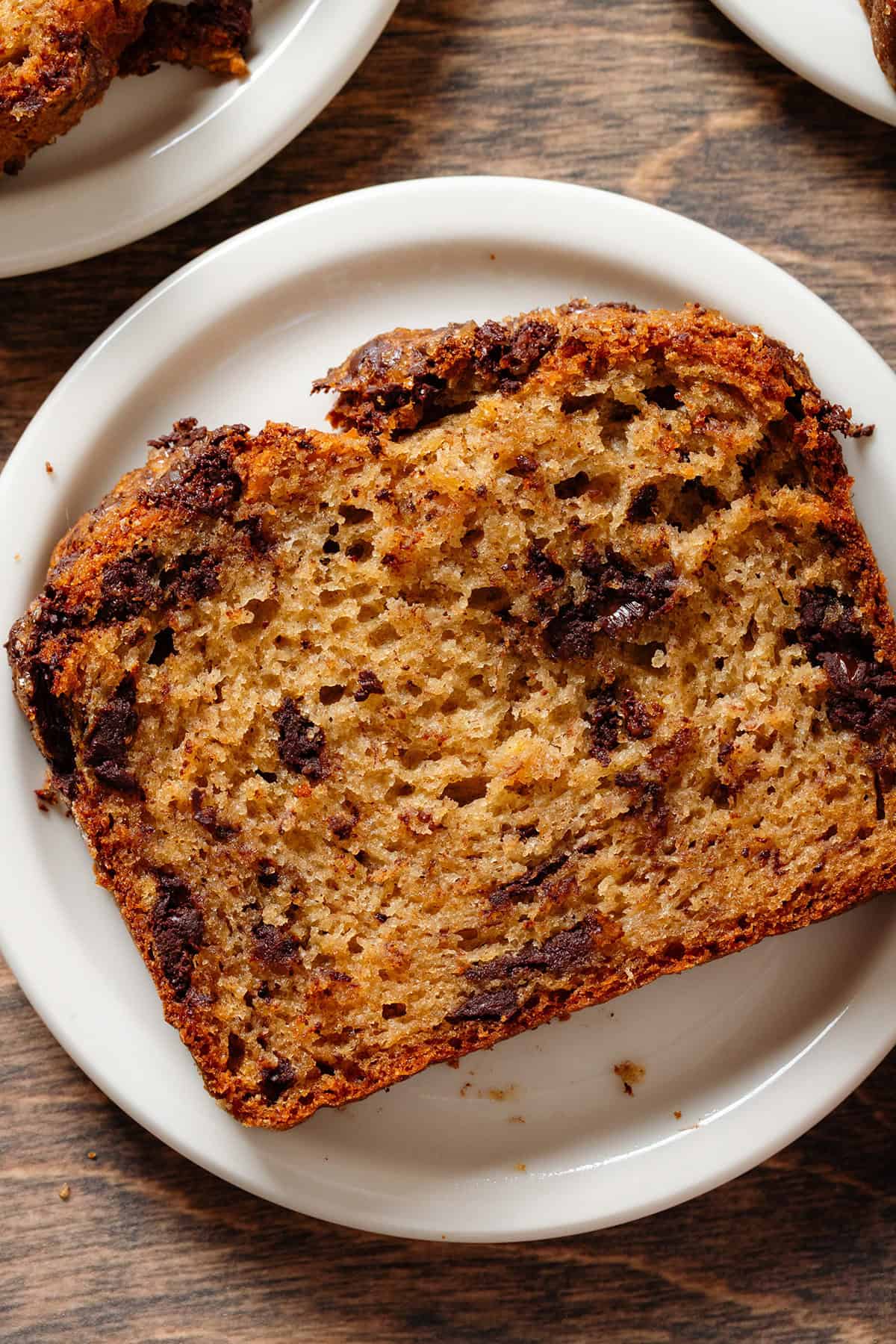 A slice of banana bread with chocolate chunks on a small white plate on a wooden background with more plates next to it.