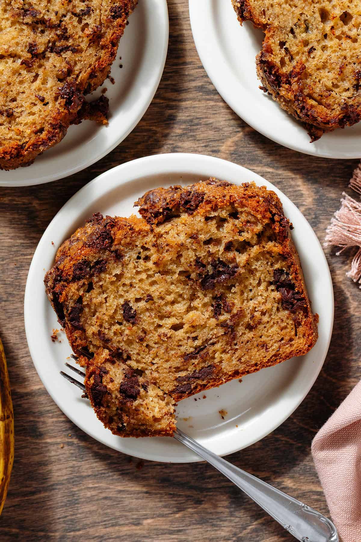 3 chocolate chunk banana bread slices on small white plates on a wooden background with a fork cutting off the corner of the slice in the middle.