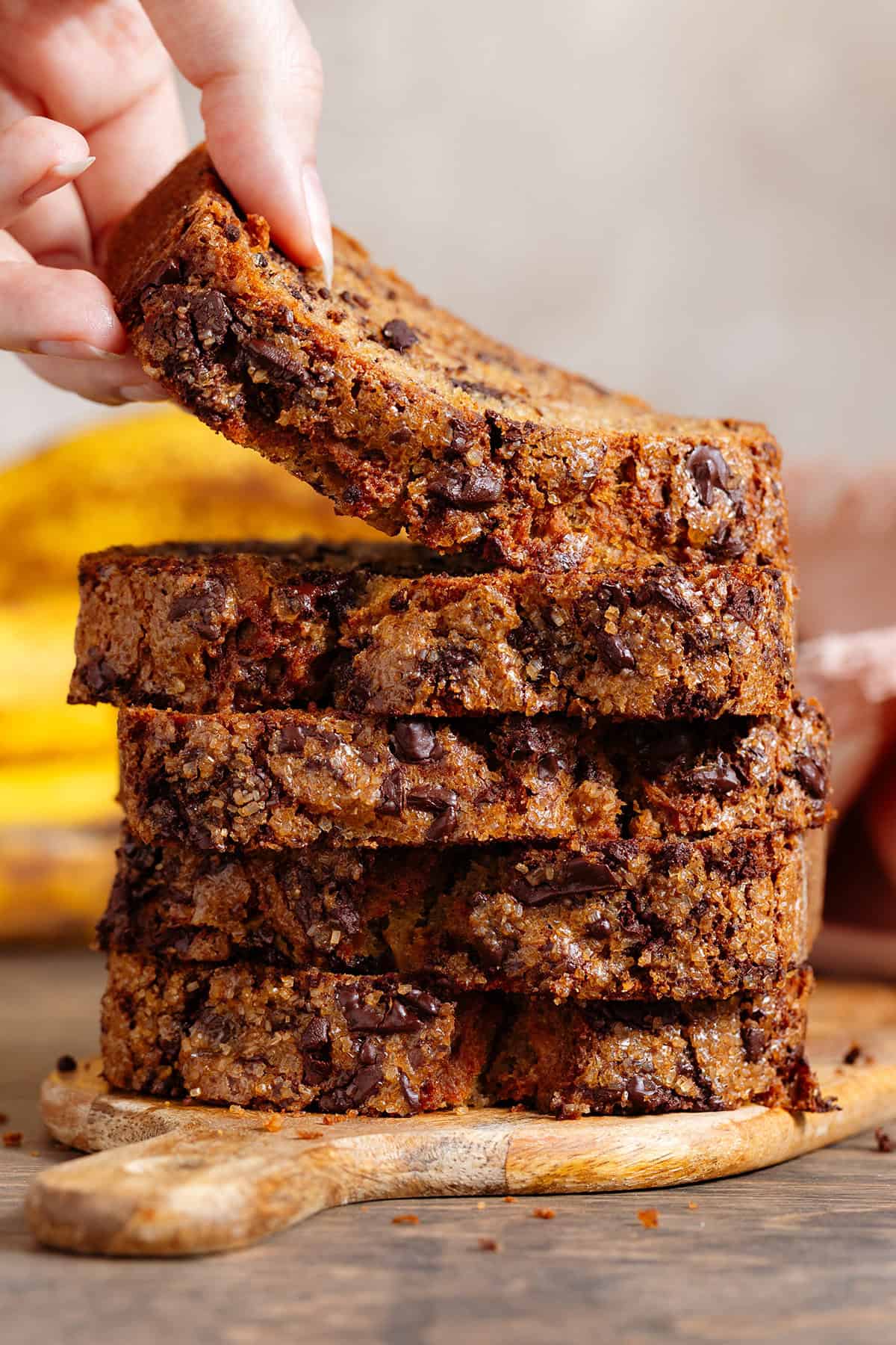 A stack of 5 chocolate chunk banana bread slices on a small wooden cutting board with a hand grabbing the slice on top.