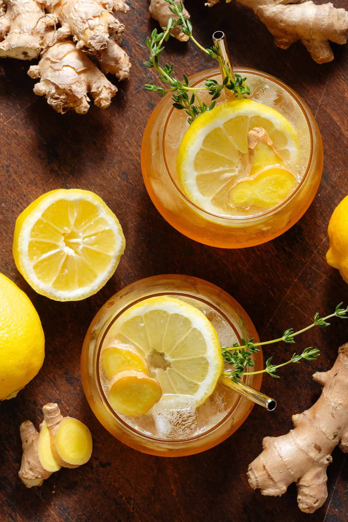 Sparkling ginger bourbon cocktail in two short glasses with ice topped with a slice of lemon, fresh ginger, fresh thyme, and a gold metal straw in each shot from above.