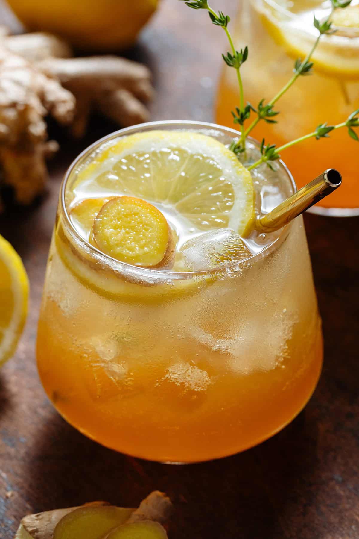 Sparkling ginger bourbon cocktail in a short glass with ice topped with a slice of lemon, fresh ginger, fresh thyme, and a gold metal straw.