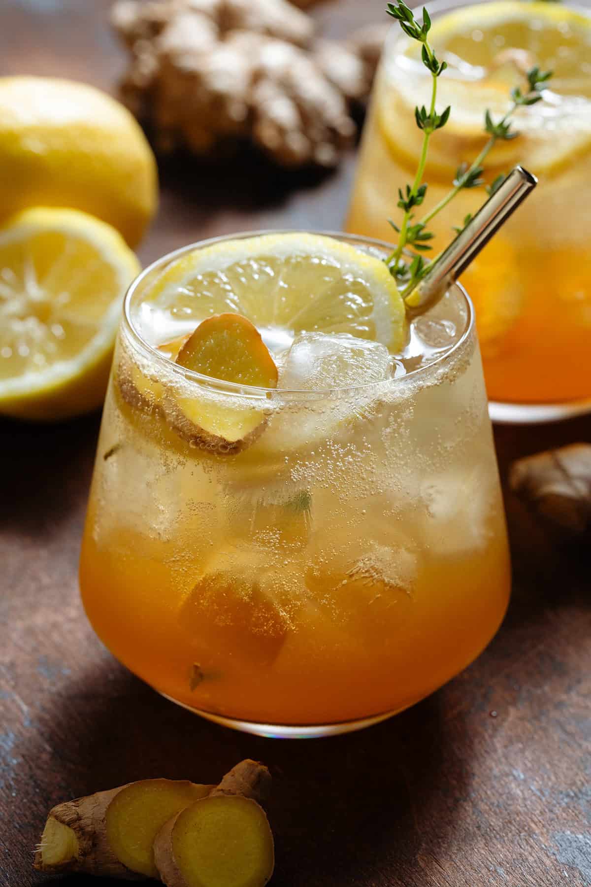 Sparkling ginger bourbon cocktail in a short glass with ice topped with a slice of lemon, fresh ginger, fresh thyme, and a gold metal straw.