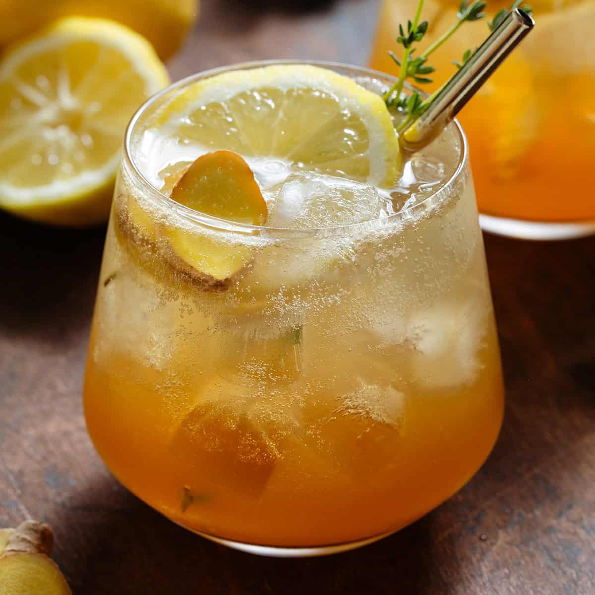 Sparkling ginger bourbon cocktail in a short glass with ice topped with a slice of lemon, fresh ginger, fresh thyme, and a gold metal straw.