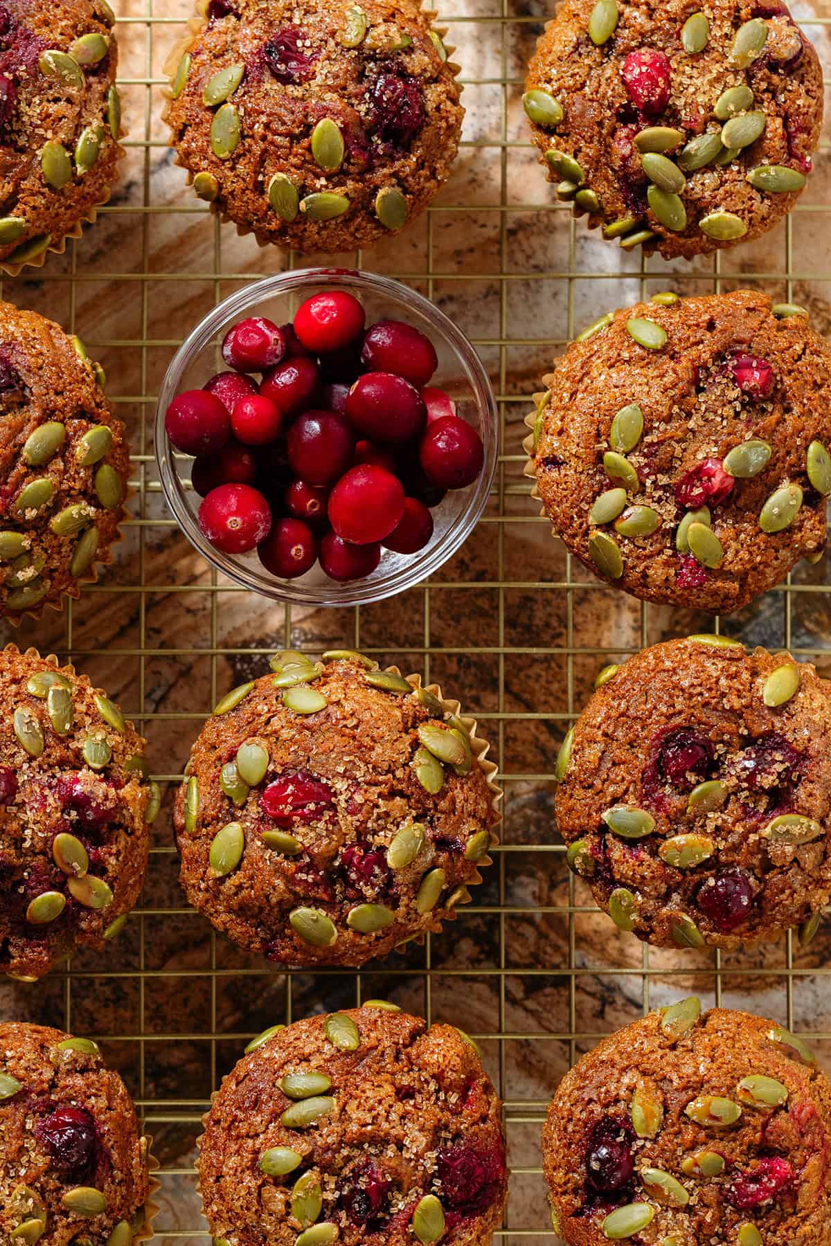 Pumpkin cranberry muffins on a golden cooling rack with a small bowl of fresh cranberries in the top left.