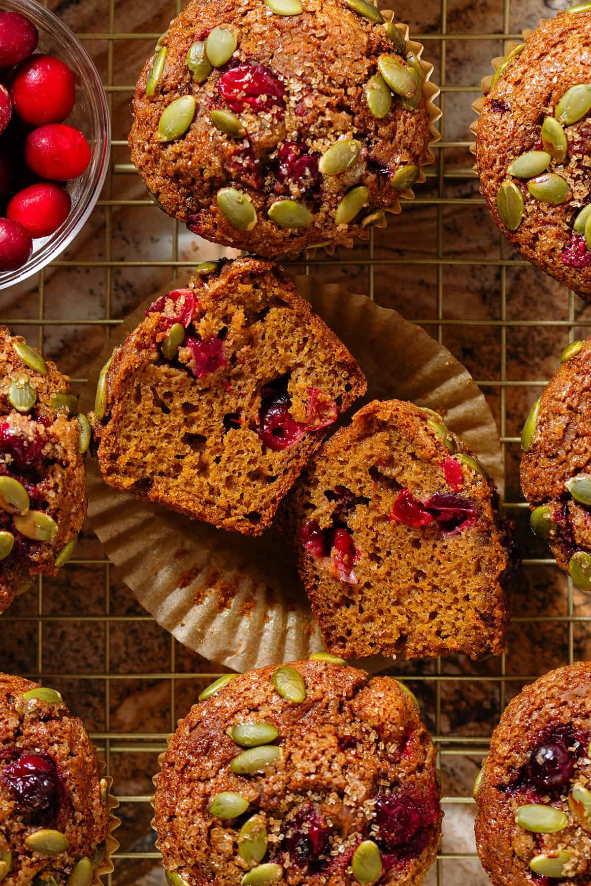 Pumpkin cranberry muffins on a golden cooling rack with the middle muffin sliced in half to show the fluffy inside with juicy cranberries and with a small bowl of fresh cranberries in the top left.