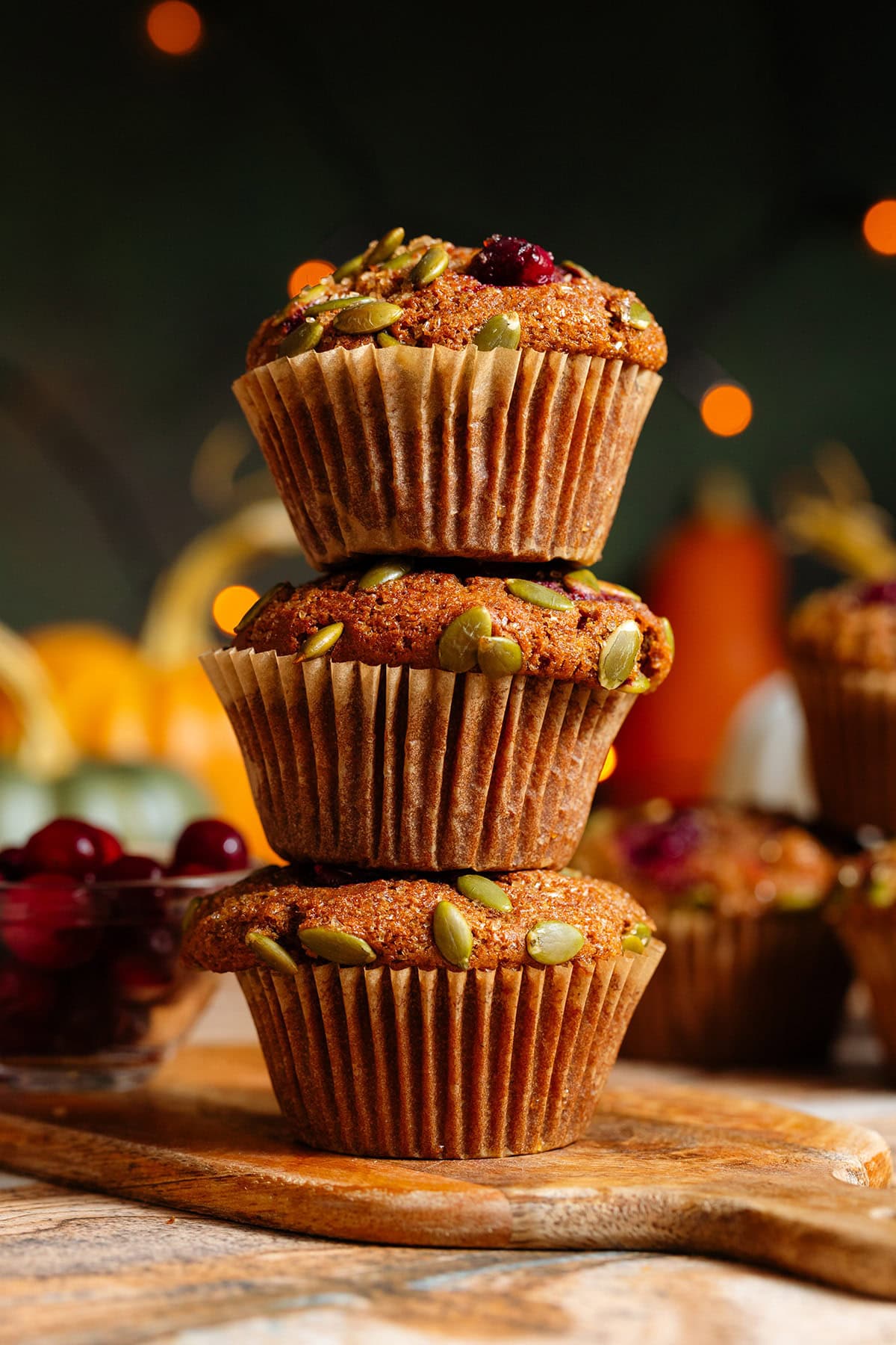 Two pumpkin muffins with cranberries and pumpkin seeds stacked on top of each other on a small wooden cutting board.