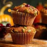 Two pumpkin muffins with cranberries and pumpkin seeds stacked on top of each other on a small wooden cutting board.