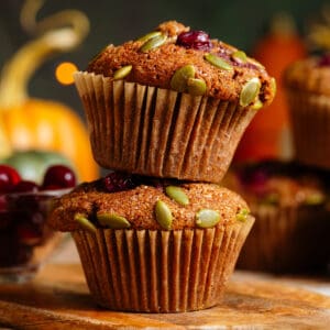 Two pumpkin muffins with cranberries and pumpkin seeds stacked on top of each other on a small wooden cutting board.