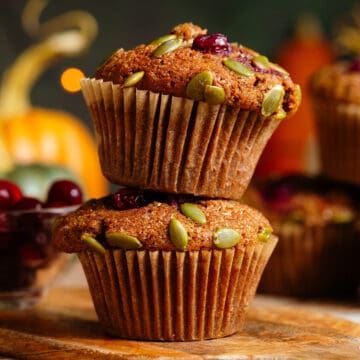 Two pumpkin muffins with cranberries and pumpkin seeds stacked on top of each other on a small wooden cutting board.