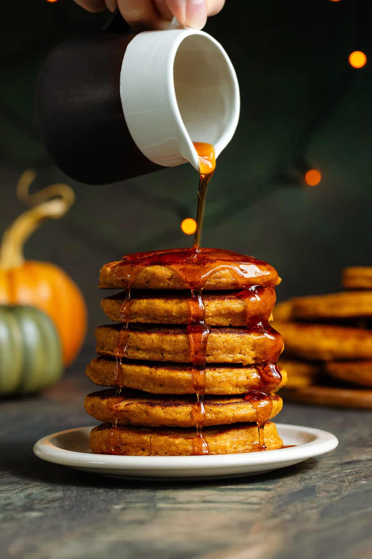 A stack of small pumpkin pancakes on a small beige plate being drizzled with maple syrup.